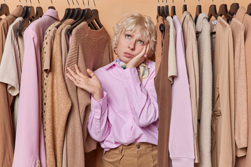 Attractive young woman dressed in fashionable outfit stands among collection of hangers filled with clothes ponders what to wear for business meeting with companions has keen sense of fashion