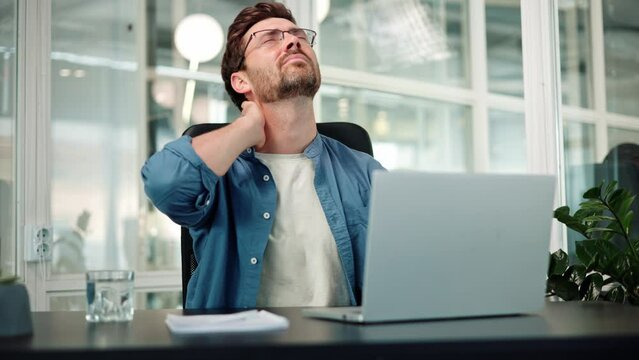 Pain in neck. Closeup of bearded man in office workplace, man experiences severe pain in neck, touches his neck with hand and makes constrained head movements. Hard work at a table with a laptop.
