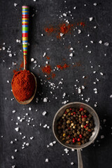 Two old spoons with peppercorn and red paprika powder on black background