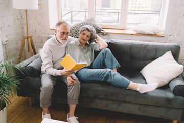 Happy elderly couple sitting on sofa and reading book together in living room