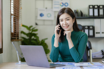Obraz premium Asian businesswoman using smartphone talking to business client and doing math on wooden table working with laptop in office, tax, accounting, finance concept
