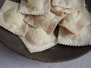 close-up view of uncooked ravioli, wholemeal homemade Italian pasta filled dusted with flour in a dark plate