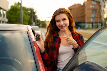 A plaid-shirted redhead woman exudes happiness as she poses next to her freshly acquired red car and pointed by finger to the key 