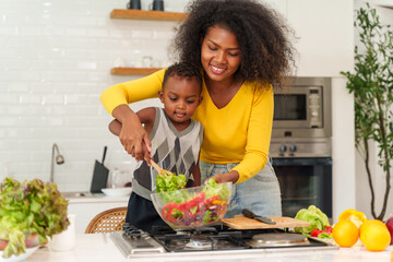 Happy African mother and son making salad while preparing food in the kitchen having fun, mother and son cooking activity concept.