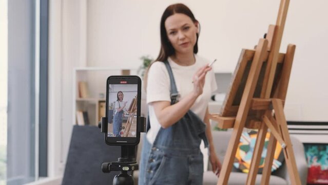 Blogger shooting video content on camera using modern smartphone and demonstrating professional skills of painting. Blur background of artist showing own technique of drawing portraits on easel.