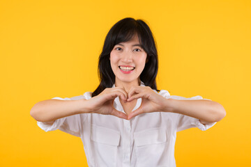 Young female healthcare. Asian woman wearing white shirt feels happy and romantic shapes heart gesture expresses tender feeling poses isolated on yellow background. People affection and care concept.