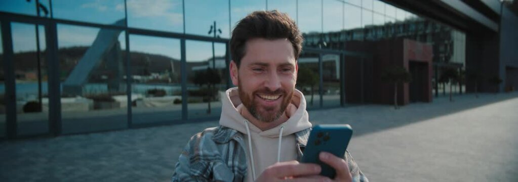 Excited young bearded man chatting on smartphone and looking around. Smiling guy in stylish clothes reading good news on background of glass modern building. Anamorphic shot