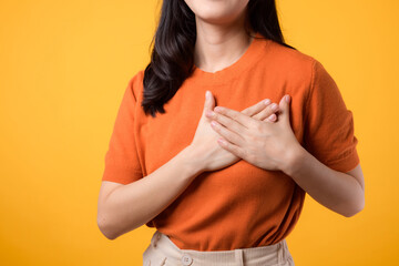 close up of young Asian woman in her 30s, wearing an orange shirt, holds hands on chest on yellow...