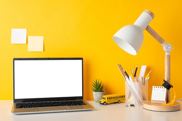 Organized study area. Side view photo of desk with pencil holder, pens, rulers, calendar, tiny academic hat, plant, open laptop, lamp, school bus toy, sticky notes against yellow wall, space for promo