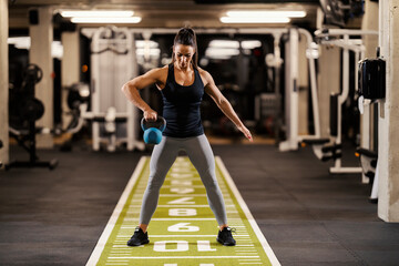 A muscular sportswoman in shape is lifting kettlebell and doing exercises for biceps in a gym.