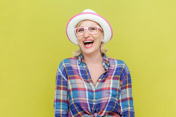 Portrait of laughing senior woman wearing checkered shirt, hat and eyeglasses being very glad, smiling with broad smile showing her perfect mood. Indoor studio shot isolated on yellow background.