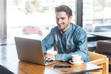 Portrait of smiling happy satisfied young man freelancer in blue jeans shirt working on laptop,...