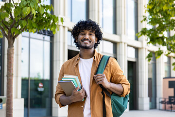 Portrait of a young smiling male Indian student standing outside the university with books in his...