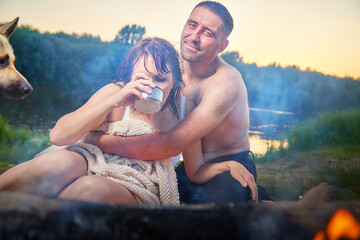 Happy wet couple relaxin, having fun and hugs with big dog near fire in camping on nature at summer sunny evening in sunset. Family or lovers have date and rest outdoor. Concept of love