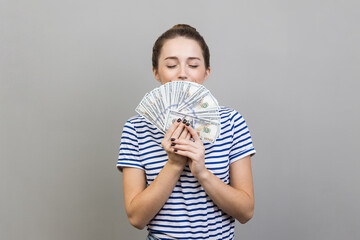 Portrait of rich satisfied woman wearing striped T-shirt holding big fan of money, smelling dollar banknotes with pleasure. Indoor studio shot isolated on gray background.