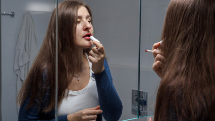 Young brunette woman looking in mirror and painting lips with red lipstick