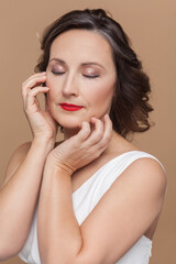 Closeup portrait of calm attractive beautiful middle aged woman with wavy hair and makeup, keeps eyes closed, wearing white dress. Indoor studio shot isolated on light brown background.