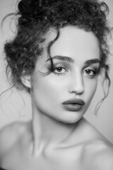 Black and white beauty portrait of young beautiful mysterious woman fashion model with collected dark curly hair, looking at camera with serious face. Indoor studio shot isolated on gray background.