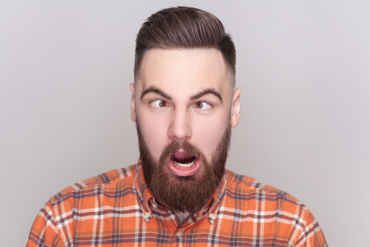 Portrait of comic positive bearded man looking cross-eyed, having fun with silly face expression, playing fool, wearing checkered shirt. Indoor studio shot isolated on gray background.