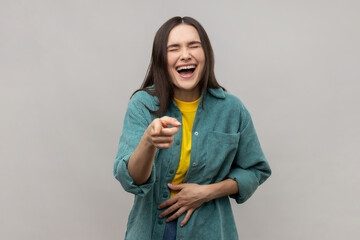 Joyful woman laughing, holding stomach, pointing to camera, taunting you, can't stop hysterical laughter, wearing casual style jacket. Indoor studio shot isolated on gray background.