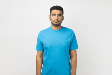 Portrait of serious strict handsome unshaven man wearing blue T- shirt standing looking at camera,...