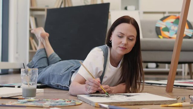 Casually dressed brunette painting picture while lying on stomach on laminate flooring at home. Smiling female artist making artwork and spending free time with paintbrush.