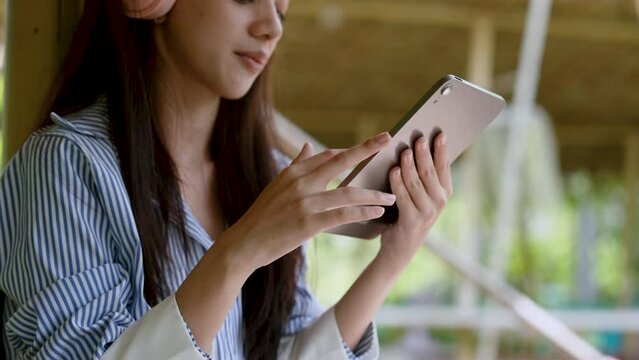 Close-up of hands of beautiful Asian Holding tablet to listen music with headphones on head. Woman uses her finger to scroll tablet to find song, smiling lightly rocking because she feels at ease.
