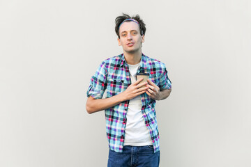 Smiling handsome man standing holding takeaway coffee in front of chest, looking at camera, love coffee, wearing blue checkered shirt and headband. Indoor studio shot isolated on gray background.