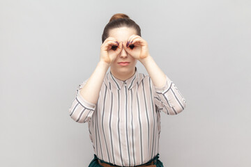 Portrait of funny positive young adult woman wearing striped shirt standing with binocular gesture, looking at something interesting. Indoor studio shot isolated on gray background.