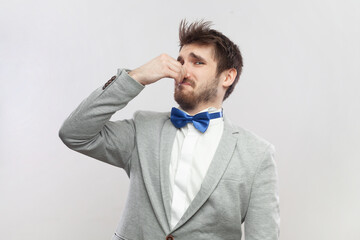 Portrait of confused handsome bearded man feels disgust smell, pinching his nose, awful aromat, wearing grey suit and blue bow tie. Indoor studio shot isolated on gray background.