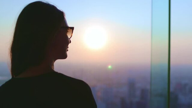 Tourist woman watching sunset over cityscape. Girl silhouette in glasses looking in window, colorful sky, city buildings, aerial view. Baiyoke Tower Sky, Thailand. Travel, tourism, slow motion 4K