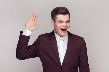 Portrait of friendly handsome young man keeps hand raise smiles sincerely expresses positive emotions, wearing violet suit and white shirt. Indoor studio shot isolated on grey background.