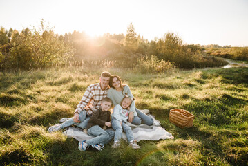 Mother, father, daughter, son sitting on blanket, hugging in grass in field at sunset. Family holiday outdoors. Children embrace parents. Happy young family walking spending time together in nature.