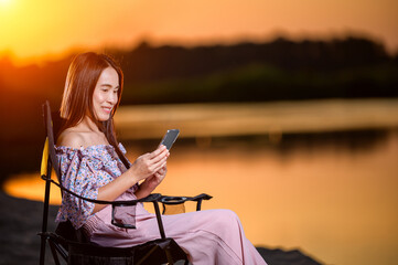 Close up Asian woman sitting on a black chair looking at smartphone on the beach during sunset