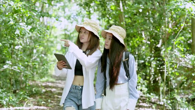 Two Asian teenage girls are best friends, holding tablet in hand, raising it open map for walk-in forest study nature this forest, two scolds each other decide which way walk.