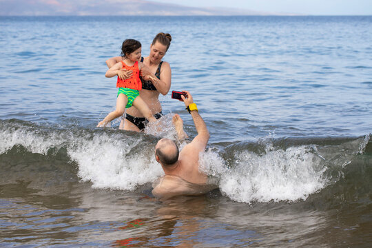 Family playing in the surf on Kamaole Beach with mother holding daughter while walking on a wave and father lying in the water taking pictures; Maui, Hawaii, United States of America