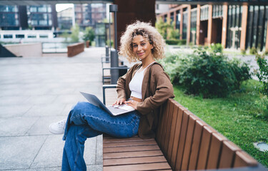 Smiling woman with laptop working remotely on bench and looking at camera