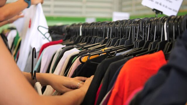 Young woman choosing t-shirt from hanger in second hand clothing store, 4K consumerism reduse reuse recycle concept footage. Chiang Mai, Thailand.