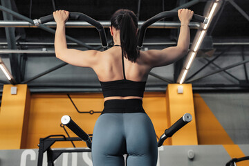 Fototapeta na wymiar Rear view of sporty woman workout pulling up on bar in fitness gym. Lady exercising pulls herself in modern sports center, working out. Healthy lifestyle and sport training concept. Copy ad text space