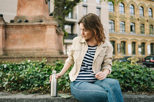 Young Woman Sitting in a City Scene, Placing Her Metal Bottle Next to Her