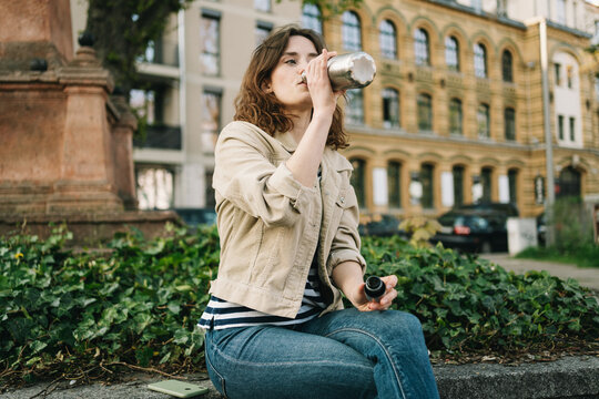 Young Woman Sitting in a City Scene, Drinking from a Metal Bottle