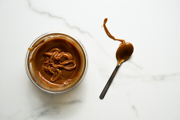Salted Caramel sauce in glass bowl on white marble background. Next to it lies a spoon soiled in caramel. Butter, sugar with cream and salt. Top view food photo.