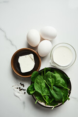 Close up of iingredients for healthy spinach and feta omelette. Eggs with heavy cream, Fresh baby spinach leaves in the black ceramic bowl, white marble background. Top view food photo
