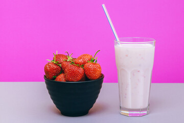 Black bowl with fresh strawberries and cold milkshake with chopped fruit in glass on gray and pink background.