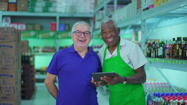 Two senior employees of Grocery store posing for camera smiling inside small business while holding tablet. Older diverse managers of supermarket