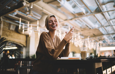 Portrait of joyful female blogger holding modern cellular device and looking at camera during weekend leisure in cafe interior, millennial hipster girl with smartphone posing while phoning