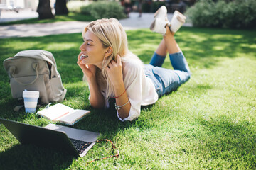 Carefree female freelancer lying at grass and smiling during positive cellphone conversation with...
