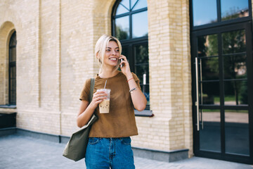 Cheerful tourist holding takeaway coffee cup and communicate during international mobile conversation for discussing vacations, roaming calling and talking via smartphone technology application