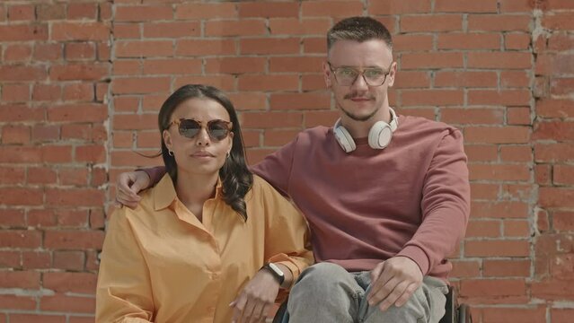 Portrait of young Caucasian wheelchaired man with his attractive Biracial girlfriend posing for camera against red brick wall outdoors