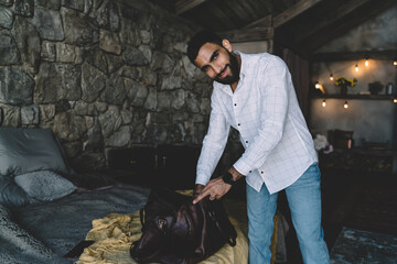 Delighted man unpacking luggage near bed